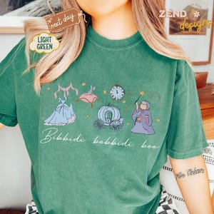May include: A light green t-shirt featuring a whimsical illustration of Cinderella's dress, carriage, and fairy godmother. The words "Bibbidi Bobbidi Boo" are written in cursive below the images. The shirt has a relaxed fit.