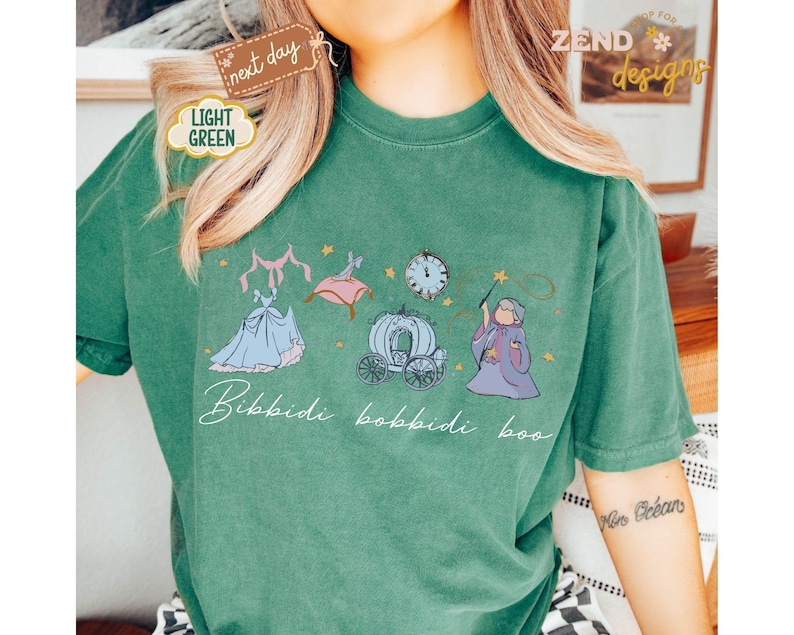 May include: A light green t-shirt featuring a whimsical illustration of Cinderella's dress, carriage, and fairy godmother. The words "Bibbidi Bobbidi Boo" are written in cursive below the images. The shirt has a relaxed fit.