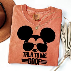 Puede incluir: Una camiseta de manga corta color terracota con un gr&aacute;fico negro de la silueta de un rat&oacute;n con gafas de sol. El texto debajo del gr&aacute;fico dice TALK TO ME GOOF. La camiseta tiene cuello redondo y una etiqueta de la marca Comfort Colors.