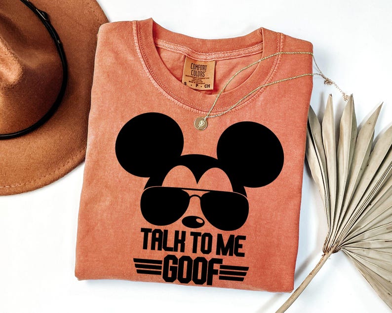 Puede incluir: Una camiseta de manga corta color terracota con un gr&aacute;fico negro de la silueta de un rat&oacute;n con gafas de sol. El texto debajo del gr&aacute;fico dice TALK TO ME GOOF. La camiseta tiene cuello redondo y una etiqueta de la marca Comfort Colors.