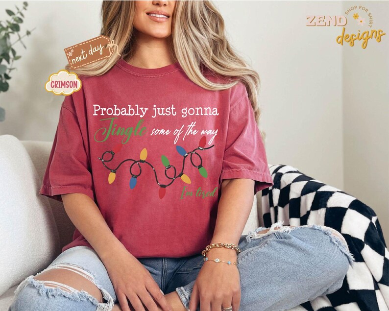 Pode incluir: Camiseta vermelha carmesim com o texto "Probably just gonna Jingle some of the way" e uma sequ&ecirc;ncia de luzes de Natal. A camisa tamb&eacute;m tem o texto "I'm tired". A camisa faz parte da cole&ccedil;&atilde;o "Zend Designs".