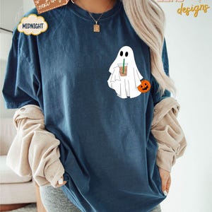 Pu&ograve; includere: Una maglietta blu con un fantasma bianco che tiene una tazza di caff&egrave; e una piccola zucca arancione. Il testo "ZEND designs" &egrave; nell'angolo in alto a destra dell'immagine.