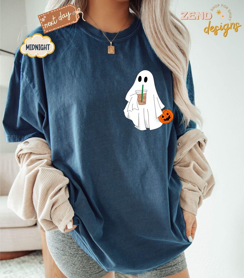 Pu&ograve; includere: Una maglietta blu con un fantasma bianco che tiene una tazza di caff&egrave; e una piccola zucca arancione. Il testo "ZEND designs" &egrave; nell'angolo in alto a destra dell'immagine.