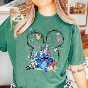 May include: A light green t-shirt featuring a graphic of Stitch from Lilo & Stitch in front of a castle. The design includes Mickey Mouse ears, fireworks, and the word "Stitch" in a script font. The shirt has a relaxed fit.