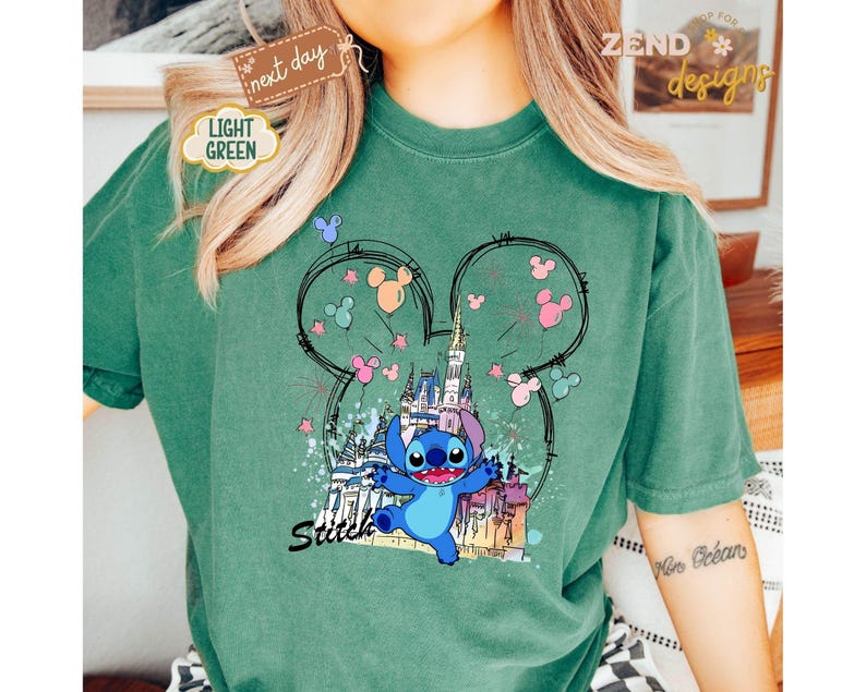 May include: A light green t-shirt featuring a graphic of Stitch from Lilo & Stitch in front of a castle. The design includes Mickey Mouse ears, fireworks, and the word "Stitch" in a script font. The shirt has a relaxed fit.