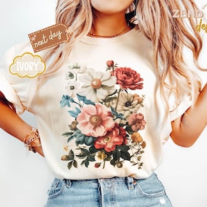 Puede incluir: Camiseta de color marfil con un dise&ntilde;o de ramo floral en tonos rosa, rojo, blanco y azul. La camiseta tiene mangas cortas y cuello redondo. Las palabras "next day" y "IVORY" est&aacute;n impresas en la camiseta.