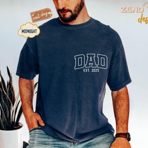 May include: A navy blue t-shirt with white text that reads "DAD EST. 2025".