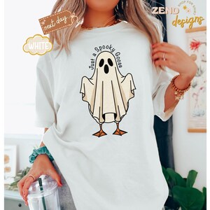 May include: White t-shirt featuring a cartoon ghost goose design with the text "Just a Spooky Goose". The ghost is white with black eyes and is standing on orange feet. The shirt has a relaxed fit.