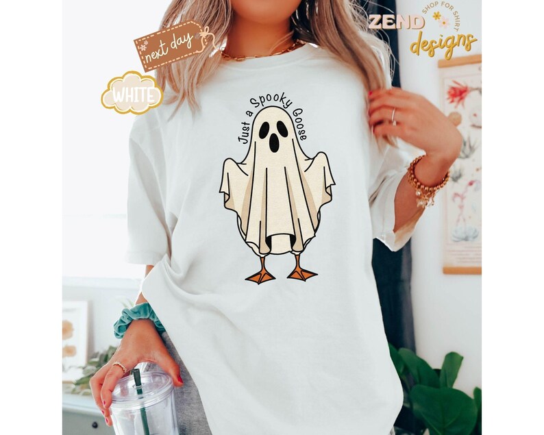 May include: White t-shirt featuring a cartoon ghost goose design with the text "Just a Spooky Goose". The ghost is white with black eyes and is standing on orange feet. The shirt has a relaxed fit.