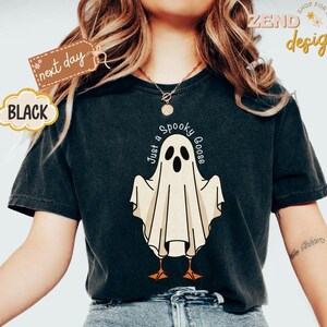 May include: Black t-shirt featuring a cartoon ghost with the text "Just a Spooky Goose". The ghost has a white body, black eyes, and orange feet. The shirt also has a "next day" tag and the word "BLACK".