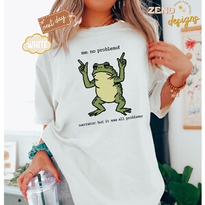 Puede incluir: Camiseta blanca con un gr&aacute;fico de rana verde. La rana est&aacute; gesticulando con las manos, y el texto dice "me: no problemo!" y "narrator: but it was all problemo". La camiseta tambi&eacute;n tiene una etiqueta "next day" y "white".