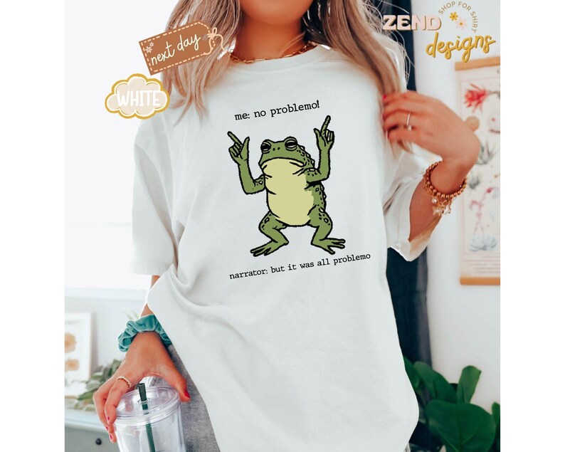 Puede incluir: Camiseta blanca con un gr&aacute;fico de rana verde. La rana est&aacute; gesticulando con las manos, y el texto dice "me: no problemo!" y "narrator: but it was all problemo". La camiseta tambi&eacute;n tiene una etiqueta "next day" y "white".