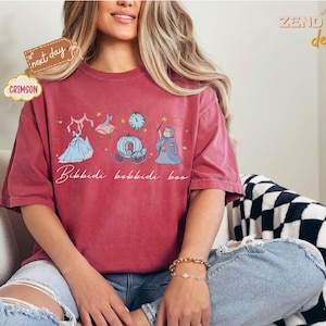 May include: Crimson-colored t-shirt featuring a whimsical design with a dress, a carriage, a clock, and a fairy godmother illustration. The phrase "Bibbidi Bobbidi Boo" is written in cursive below the illustrations. The shirt is a comfortable, oversized fit.
