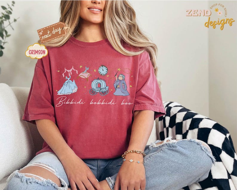 May include: Crimson-colored t-shirt featuring a whimsical design with a dress, a carriage, a clock, and a fairy godmother illustration. The phrase "Bibbidi Bobbidi Boo" is written in cursive below the illustrations. The shirt is a comfortable, oversized fit.