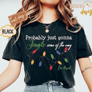 Pode incluir: Camiseta preta com a frase "Probably just gonna Jingle some of the way" em escrita branca e verde, com uma sequ&ecirc;ncia de luzes de Natal coloridas. A frase "I'm tired" est&aacute; abaixo das luzes.