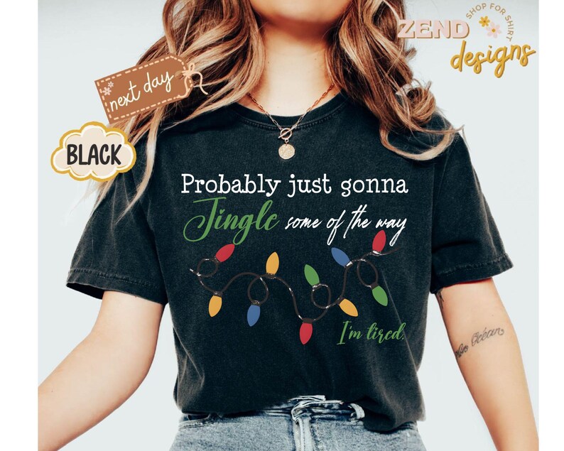 Pode incluir: Camiseta preta com a frase "Probably just gonna Jingle some of the way" em escrita branca e verde, com uma sequ&ecirc;ncia de luzes de Natal coloridas. A frase "I'm tired" est&aacute; abaixo das luzes.