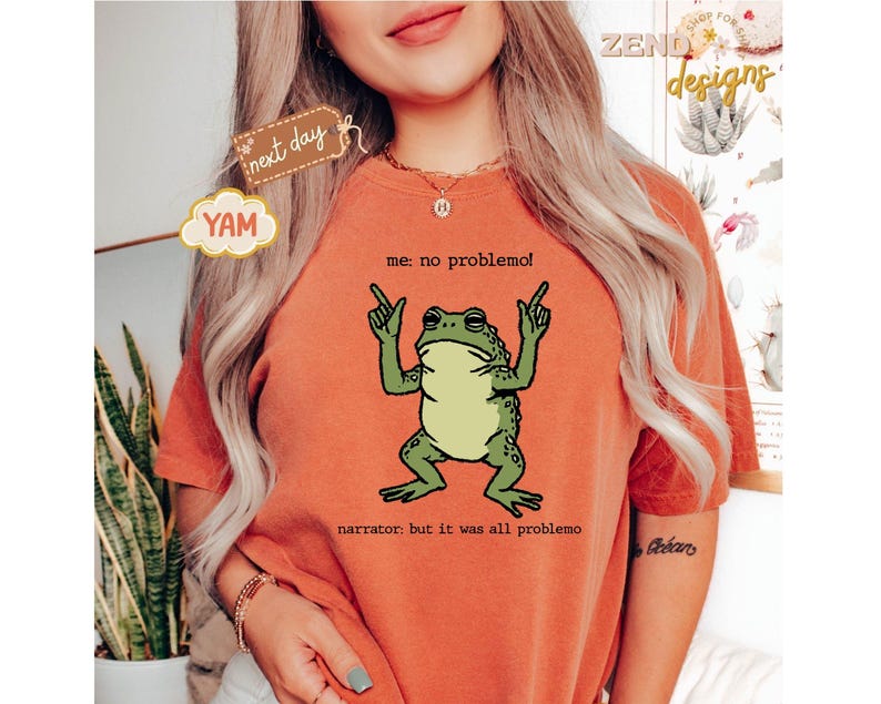 Puede incluir: Camiseta naranja con un dise&ntilde;o de rana de dibujos animados. La rana es verde y beige, con los brazos levantados, y el texto dice "me: no problemol" y "narrator: but it was all problemo". La camiseta tiene cuello redondo.