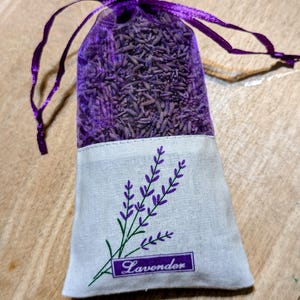 Peut inclure: Un sachet violet rempli de boutons de lavande séchés. La partie supérieure est transparente, tandis que le bas est en tissu clair avec un motif de brins de lavande et le mot "Lavender". Le sachet est noué avec un ruban violet.