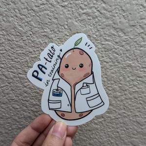 May include: A sticker of a cartoon potato wearing a white lab coat with a name tag that says "Potato Student". The potato has a green sprout on top and a happy expression. The text "PA-tato in training" is written in blue and white lettering around the potato.