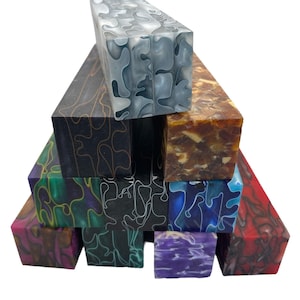 May include: A collection of colourful, patterned acrylic blanks for crafting. The blanks are in various shades of blue, green, purple, red, brown, and white. They are arranged in a pyramid shape.