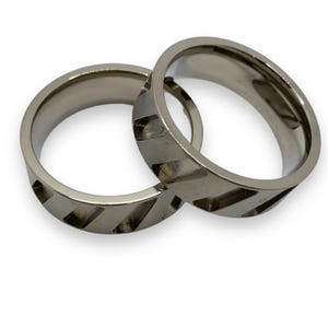 May include: Two silver-colored rings with a diagonal cut-out design. The rings are made of a shiny metal and are displayed on a white background. The rings are likely for a couple or a pair.