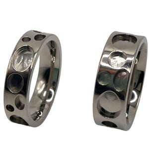 May include: Two silver rings with a unique design featuring multiple circular cutouts. The rings are smooth and shiny.
