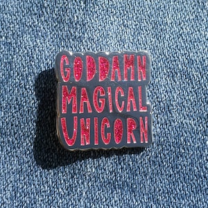 以下が含まれることがあります： 「GODDAMN MAGICAL UNICORN」とピンクのグリッター文字で書かれた黒と銀の七宝焼きピン。