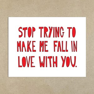 Op de afbeelding: Een witte kaart met de rode tekst: "STOP TRYING TO MAKE ME FALL IN LOVE WITH YOU." De kaart staat tegen een neutrale, getextureerde achtergrond.