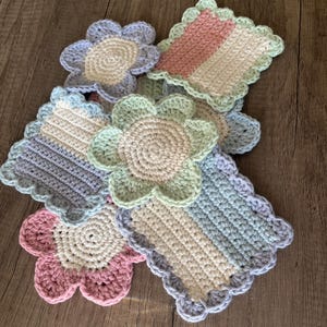 May include: Five crocheted flower-shaped coasters in pastel colours. The coasters are made of white, pink, light blue, and green yarn. They are arranged in a pile on a wooden surface.