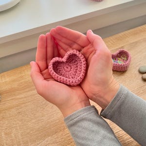 Könnte beinhalten: Eine rosa gehäkelte Schale in Herzform, die in den Händen einer Person gehalten wird. Die Schale ist klein und wäre perfekt, um Schmuck oder andere kleine Gegenstände aufzubewahren.