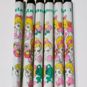 May include: A set of six white pencils with black tips. Each pencil features a cartoon illustration of a girl with yellow hair, wearing a pink dress and a pink bow. The pencils have the text "la Bebe" printed on them.
