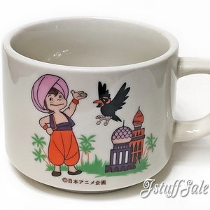 Peut inclure: Une tasse en céramique blanche avec une illustration de dessin animé d'un homme portant un turban et un pantalon orange, un oiseau noir et un bâtiment avec un dôme. Le texte "© 日本アニメ企画" est imprimé au fond de la tasse.
