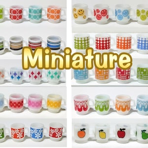 May include: A selection of miniature glass mugs featuring diverse, colourful patterns such as smiley faces, dots, stripes, and floral motifs. The word "Miniature" is displayed. Each mug has a handle.
