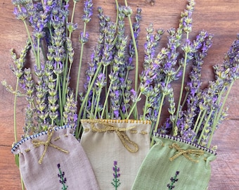 May include: Three small fabric pouches in light purple, beige, and sage green, each featuring a single embroidered lavender sprig and a gold metallic drawstring tie. The pouches are arranged among several stems of fresh purple lavender flowers.
