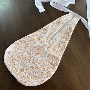 May include: White fabric apron with detailed gold floral embroidery. The apron has a long, pointed shape with a split at the top and white ties. The floral design includes detailed flowers and leaves.