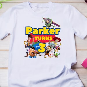 May include: A white short-sleeve t-shirt featuring a colorful graphic print of Toy Story characters including Woody, Buzz Lightyear, Jessie, and Mr. Potato Head. The text reads Parker turns 3 in bold blue and yellow lettering with a small blue frog.