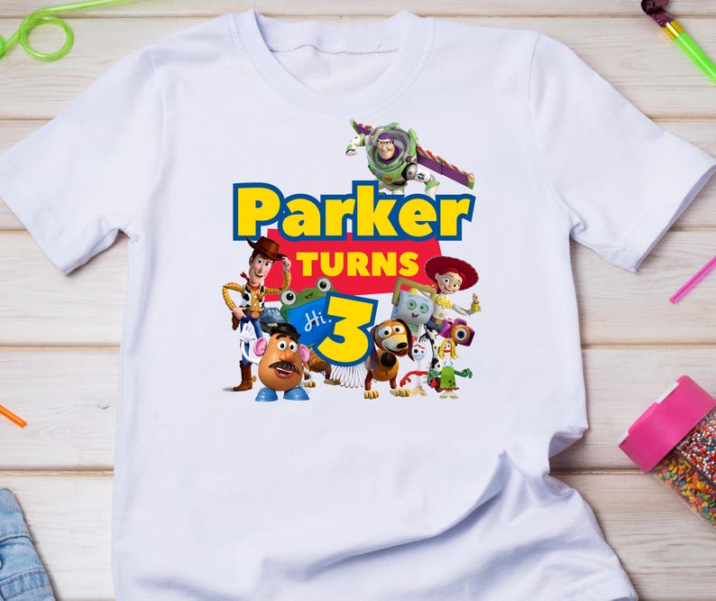 May include: A white short-sleeve t-shirt featuring a colorful graphic print of Toy Story characters including Woody, Buzz Lightyear, Jessie, and Mr. Potato Head. The text reads Parker turns 3 in bold blue and yellow lettering with a small blue frog.