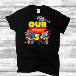 May include: A black short-sleeved t-shirt featuring a graphic print of Toy Story characters including Woody, Buzz Lightyear, Jessie, Mr. Potato Head, and others. The text "OUR STORY" appears in blue and yellow, with a large yellow number 5 below.
