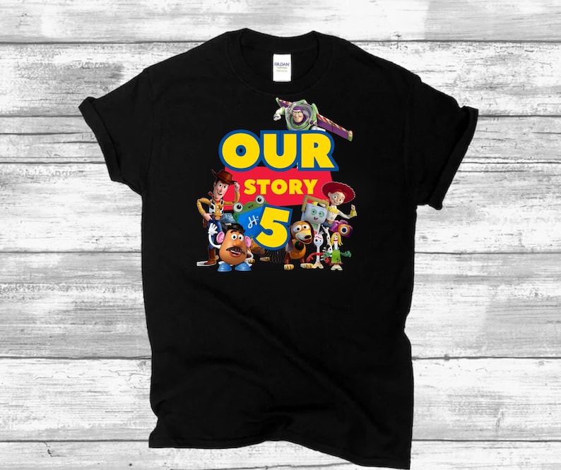May include: A black short-sleeved t-shirt featuring a graphic print of Toy Story characters including Woody, Buzz Lightyear, Jessie, Mr. Potato Head, and others. The text "OUR STORY" appears in blue and yellow, with a large yellow number 5 below.