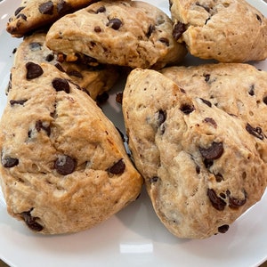 May include: A plate of six chocolate chip scones. The scones are light brown and have a slightly crispy exterior. The chocolate chips are scattered throughout the scones.