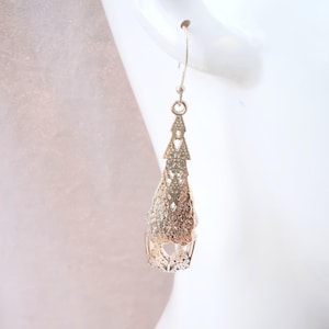 May include: A gold-toned dangle earring featuring an elongated, tapered design with intricate filigree metalwork. The earring hangs from a simple hook wire and displays a delicate, open-patterned texture throughout its entire surface structure.