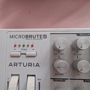 May include: A white Arturia MicroBrute SE analog synthesizer featuring a control panel with silver knobs, two white pitch and modulation wheels, and an octave selection section with five small indicator lights labeled from -2 to +2.