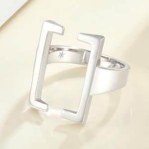 May include: A polished silver-tone ring featuring a large, open rectangular frame design that extends upward from the band. The interior of the band displays a small, engraved starburst or spiral symbol centered on the inner surface of the metal.