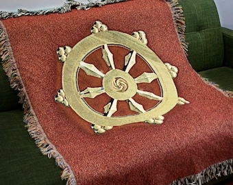 May include: A terracotta-coloured throw blanket featuring a gold and cream Dharma wheel design. The blanket has a fringed edge and is draped over a green sofa. The Dharma wheel is a symbol of Buddhism.