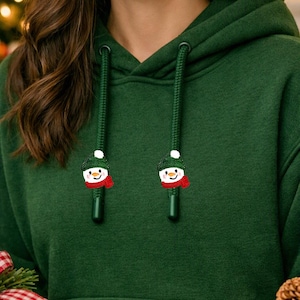 May include: A dark green hooded sweatshirt featuring two matching drawstring cords. Each cord ends in a small, embroidered snowman charm wearing a green hat and a red scarf, positioned above a front kangaroo pocket.