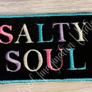 May include: Embroidered patch with the words "SALTY SOUL" in colorful letters on a black background. The patch has a turquoise border and is likely for clothing or accessories.