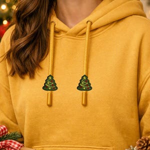 May include: A mustard yellow hooded sweatshirt featuring two matching yellow drawstrings. Each drawstring is finished with a small, embroidered green Christmas tree patch with red and yellow ornaments and a yellow star topper.
