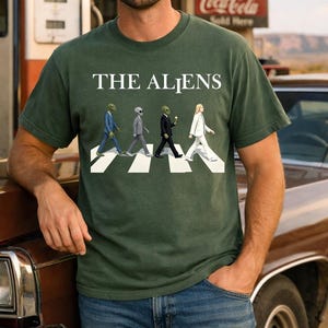 May include: A dark green short-sleeve t-shirt featuring the text THE ALIENS in white capital letters above a graphic of four extraterrestrial figures in suits walking across a white-striped crosswalk, mimicking a famous album cover design.