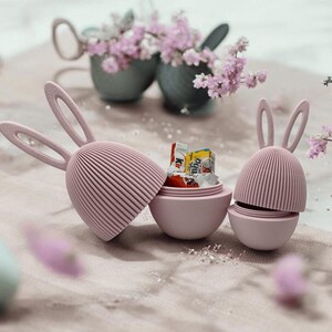 Può includere: Tre contenitori per uova di Pasqua a forma di coniglio rosa, uno dei quali è aperto e riempito con piccole barrette di cioccolato. I contenitori hanno una texture a coste e sono su una superficie bianca e soffice.