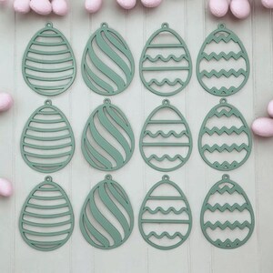 Può includere: Dodici uova di Pasqua in legno verde con diversi disegni ritagliati. Le uova sono disposte in quattro file di tre.
